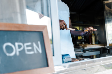 cropped view of afrian american man holding spatula while preparing burger near chalk board with open letters
