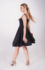 Young attractive sexy blond woman model with light hair, wearing short black dress and sandals on heels, posing and dancing in white studio