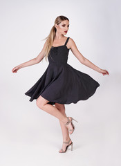 Young attractive sexy blond woman model with light hair, wearing short black dress and sandals on heels, posing and dancing in white studio