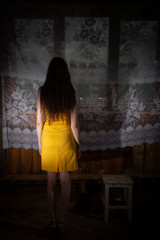 A girl in a yellow dress stands and looks out the window. Photo from the back. Wooden unfinished...