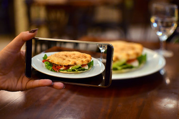 Woman taking photo of vegetarian sandwich with mozzarella, gorgonzola, mushrooms, lettuce, tomato and bread with sesame