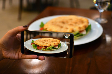 Woman taking photo of vegetarian sandwich with mozzarella, gorgonzola, mushrooms, lettuce, tomato and bread with sesame
