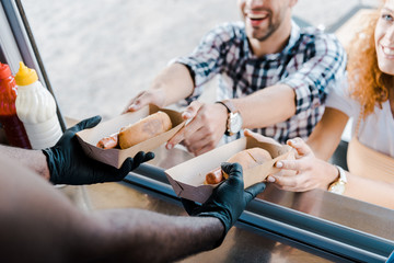 cropped view of african american man holding carton plates with hot dogs near happy customers