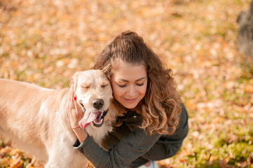 Portrait of a curly young woman hugging her golden retriever dog in the autumn park. yellow leaves background. Young curly woman sitting with her dog in autumn leaves