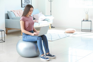 Young woman with laptop sitting on fitball at home