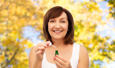 beauty, skincare and cosmetics concept - smiling senior woman with dropper and bottle of serum over natural autumn background