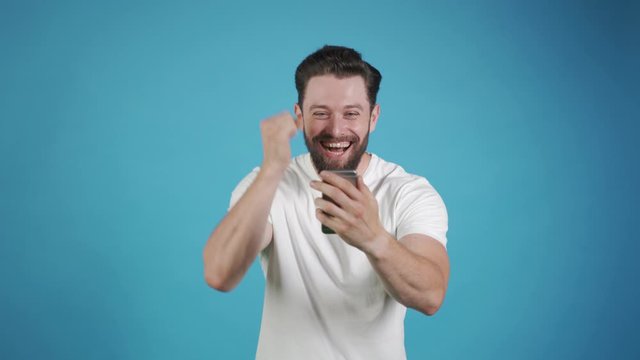 Happy man holding smartphone and celebrating his success over blue background. Victory, win, success. Best result.