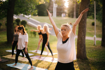 Obraz premium Group of young women do yoga in city park on summer sunny morning under guidance of instructor
