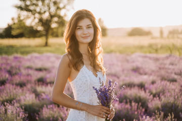 Beautiful Young Woman Portrait in Lavender Field. Attractive Brunette Girl with Long Wavy Hair Looking at Camera. Caucasian Model Posing with Bunch of Purple Flowers. Blooming Meadow Background