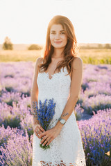 Young Woman Stand on Lavender Field Background. Caucasian Girl with Pretty Smile Looking at Camera. Attractive Model in White Dress Hold Purple Bouquet. Blooming Flower Meadow on Sunset