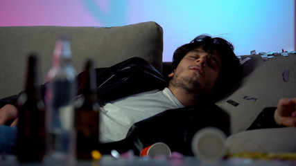 Drunk man sleeping on sofa after party at home, empty bottles on table, addict