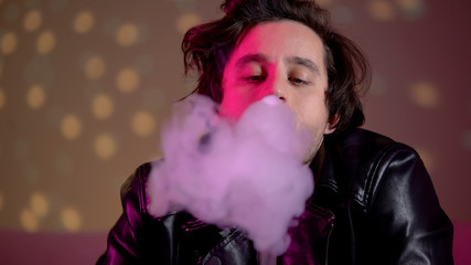 Young man exhaling e-cigarette smoke at camera, vaping at rave party in club