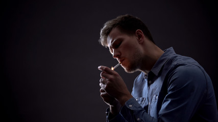 Young man lighting cigarette in dark room, unhealthy habit, harmful effect