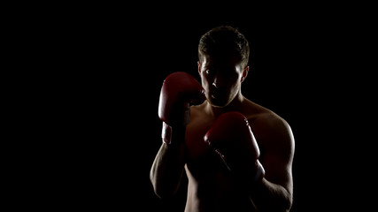 Strong man in boxing gloves preparing to fight, training in gym black background