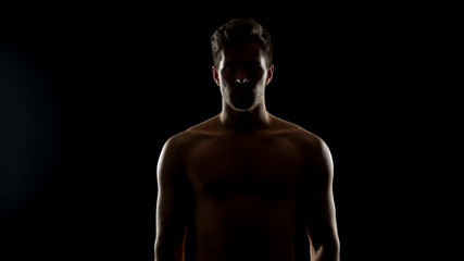 Shadowed topless man standing against black background, hopelessness concept