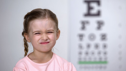 Cute girl screwing up her eyes, trying to see letters on table for vision test