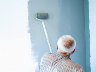 An elderly man paints a wall with a roller. Senior man makes repairs. painting the walls with a roller. old outfit. gray wind on a white wall. home repair
