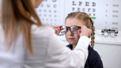 Optician putting glasses on schoolgirl, checking vision and recommending glasses
