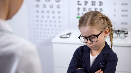 Female doctor putting glasses on disgruntled girl, child feels insecure, upset