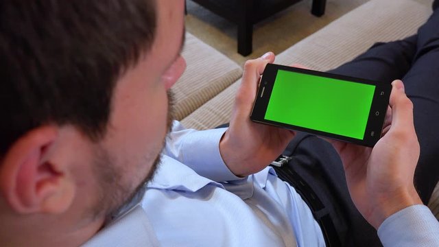 A man sits on a couch in an apartment and looks at a smartphone with a green screen in horizontal position - closeup