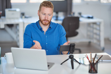 Fototapeta premium Young Caucasian employee having a video call over smart phone while sitting in office. On desk is laptop. Start up business concept.