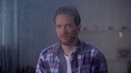 Lonely man sitting behind rainy window and looking at camera, depression concept