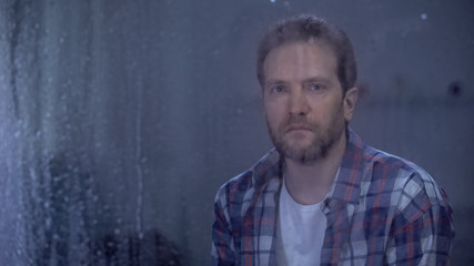 Obraz na płótnie Canvas Lonely man looking at camera through window on rainy day, depression problems