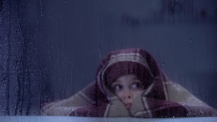 Scared little girl covered with blanket frightened by thunder on rainy day