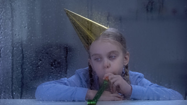 Upset girl in birthday hat blowing party horn on rainy day, celebrating alone