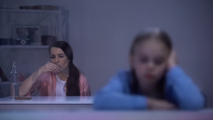 Sad little girl sitting on rainy day, addicted mother drinking alcohol in room