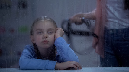 Mother with belt standing behind little girl on rainy window, assault in family