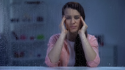 Exhausted woman suffering from head ache behind rainy window, migraine disorder
