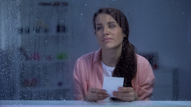 Sad woman with photo feeling sad behind rainy window, remembering first love