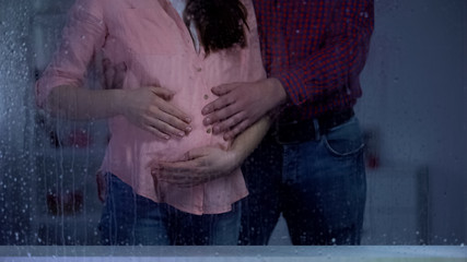 Couple holding pregnant woman tummy behind rainy window, baby expectation