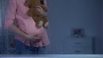 Pregnant woman hugging teddy bear behind rainy window, baby expectation, care