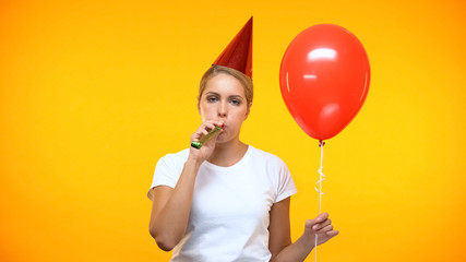 Sad adult woman blowing noisemaker holding balloon, bad mood at birthday party