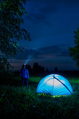 Male tourist with a flashlight looks at the night sky. Near his blue lighted tent in the forest.