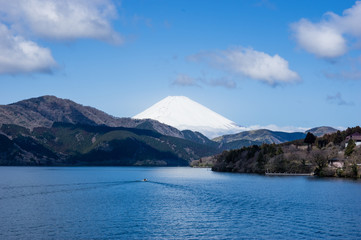 Mount Fuji in Japan covered by snow seen across a large lake. Small boat leaving a triangle form wake as if mirroring the shape of the mountain. Blue sky, white clouds, spring in coastal forests