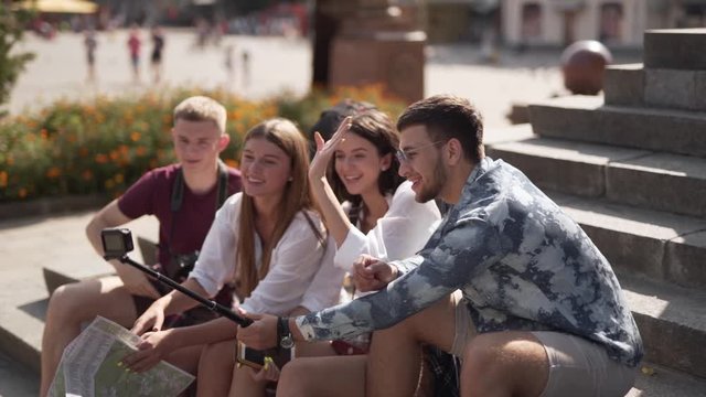 Four young friends are sitting on a stairs of some ancient building and making a selfie. They are tourists cause they are hoding a map of the city.