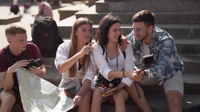 Four young friends are sitting on a stairs of some ancient building and making a selfie. They are tourists cause they are hoding a map of the city.