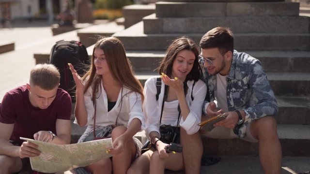 Four young friends are sitting on a stairs of some ancient building and looking at the map of the city. They are tourists and they are deciding the way where to go.