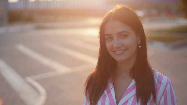 Beautiful young cheerful wooman smiling and playing with hair on the streer at sunset. Closeup portrait of positive girl outdoors in Slow motion
