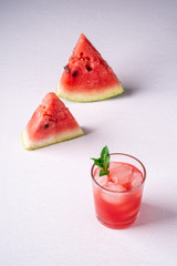Fresh cold watermelon juice with ice cubes and green mint leaf in glass drink close to two slices of watermelon on white background, copy space, angle view, selective focus