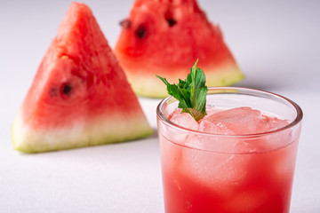Fresh cold watermelon juice with ice cubes and green mint leaf in glass drink close to two slices of watermelon on white background, copy space, angle view, selective focus