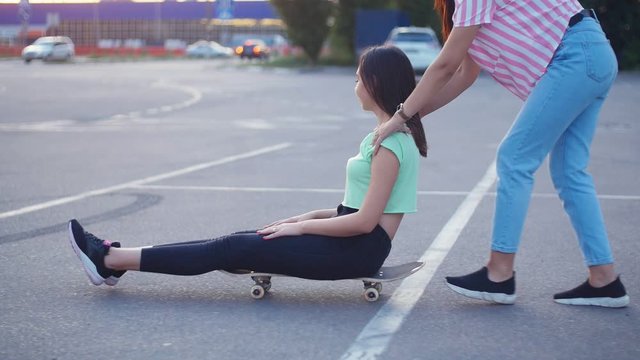 Positive girls having fun with scateboard on the parking at sunset in Slow motion. Two young cheerful women riding on the skateboard