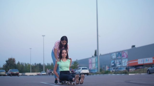 Positive girls having fun with scateboard on the parking at sunset in Slow motion. Two young cheerful women riding on the skateboard