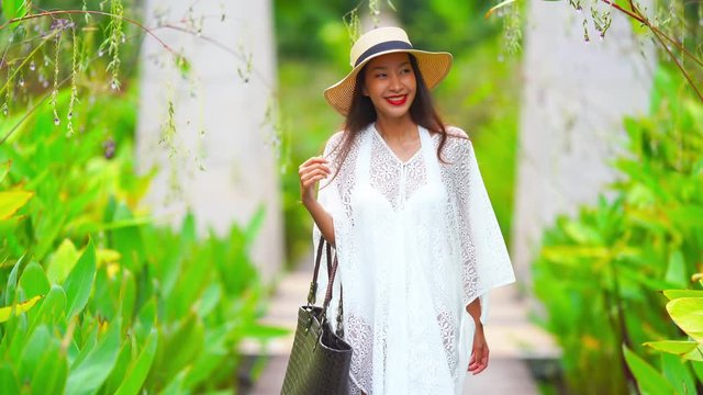 Charming asian lady wearing white dress and hat, holding bag on right hand walking towards the camera smiling along the walkway, tropical plants background.
