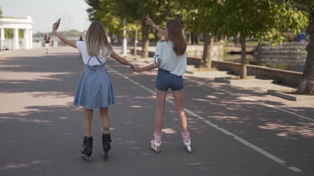 girls in rollerblades taking a selfie while skating in the park hand in hand, shot from the back