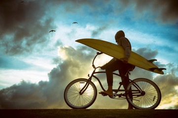 Silhouette of surfer carrying his surfboard on a beach cruiser bike in front of bright skyscape