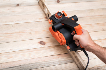 Electric planer in male hand. Processing of the workpiece on light brown wooden table.